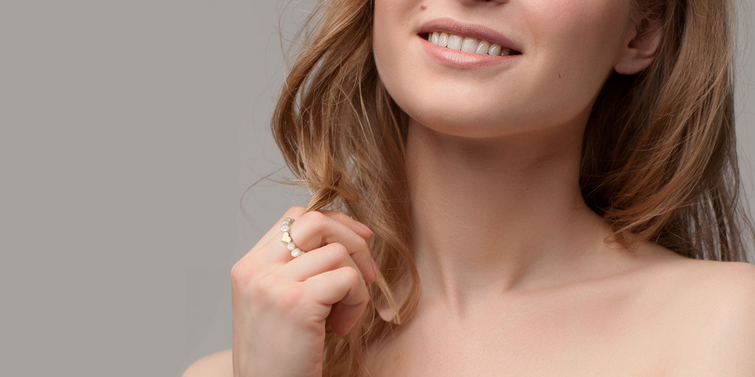 Lächelnde Frau mit braunen Haaren trägt Ring mit weißen Perlen und einem kelinen goldenen Herz