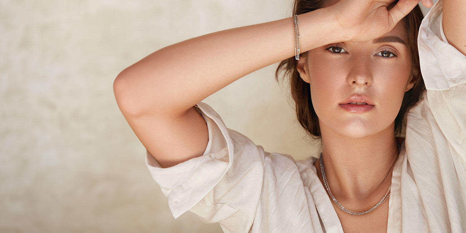 Junge Frau mit braunen Haaren in entspannter Pose in sommerlichem weißem Hemd trägt mehrreihige silberne Halskette und ein passendes silbernes Armband 